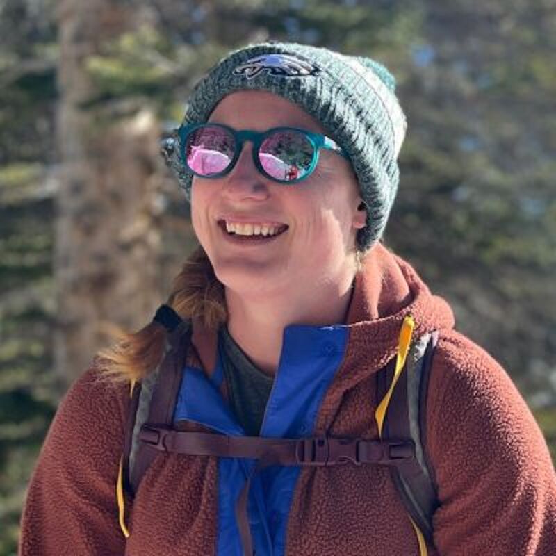 The image shows a woman wearing a beanie, sunglasses, and a jacket. She is smiling and appears to be outdoors, possibly hiking or enjoying nature. The background includes trees, suggesting a natural environment. The woman seems happy and relaxed.